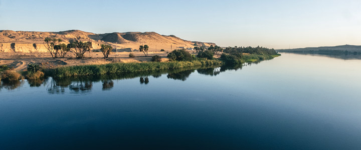 Eine Reise durch Ägypten führt entlang der Weite des Nils, wo ein schmaler Grünstreifen auf die dahinterliegende Wüste trifft. Das Ufer wirkt ruhig und weit.