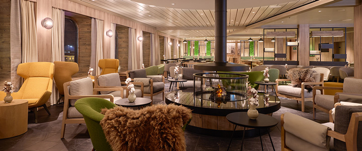 Ein Blick in die Lounge der Mein Schiff Flow zeigt eine ruhige Atmosphäre in warmen Brauntönen. Edles Licht und ein Kamin schaffen eine gemütliche, stilvolle Umgebung.