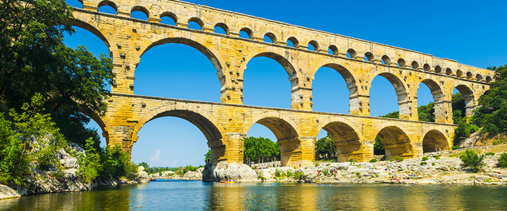 Vom Wasser aus erhebt sich der Pont du Gard monumental in den Himmel. Strahlendes Blau umgibt die alte Steinbrücke.