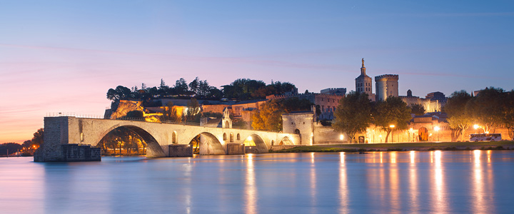 In der Abendstimmung spiegelt sich Avignon im ruhigen Fluss. Die beleuchtete Brücke und Skyline zeichnen sich klar gegen den Himmel ab.