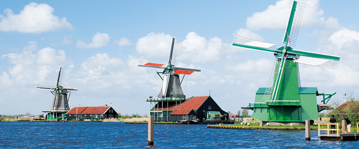 Blick über blaues Wasser auf die Windmühlen der Zaanse Schans. Grüne Wiesen und klare Luft prägen die weite Landschaft Nordhollands.
