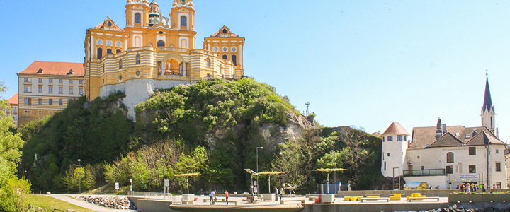 Vom Donauufer aus erhebt sich das Stift Melk eindrucksvoll in den Sommerhimmel. Goldene Fassaden leuchten über der grünen Flusslandschaft.