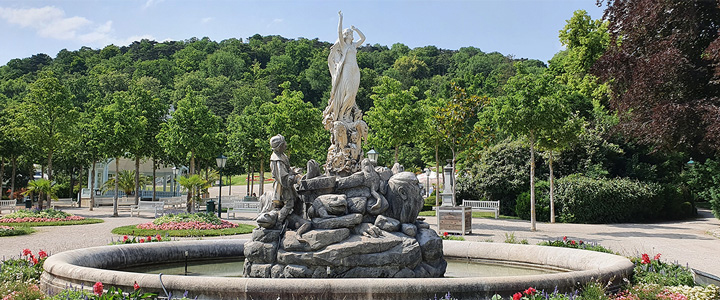 Im Kurpark Baden erhebt sich eine Skulptur neben dem Springbrunnen. Grünanlagen und ruhige Wege prägen die sommerliche Atmosphäre.