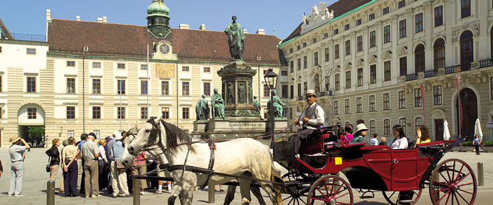 Vor der Hofburg in Wien fährt eine Kutsche mit weißen Pferden über das Kopfsteinpflaster. Die historische Fassade rahmt die Szene.