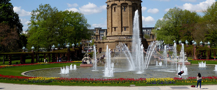 Im Vordergrund sprudelt der Springbrunnen am Wasserturm Mannheim. Dahinter erhebt sich das markante Bauwerk im klaren Stadtbild.